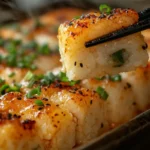 A freshly baked sushi bake in a ceramic dish, topped with furikake and drizzled with spicy mayo, with a piece being lifted using chopsticks and wrapped in a nori sheet.